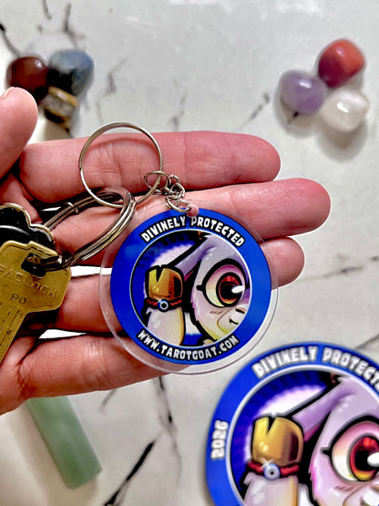 A hand holding a round acrylic keychain featuring a blue border and a cartoon Goat Goated Energy illustration with the text 'DIVINELY PROTECTED' and 'WWW.TAROTGOAT.
