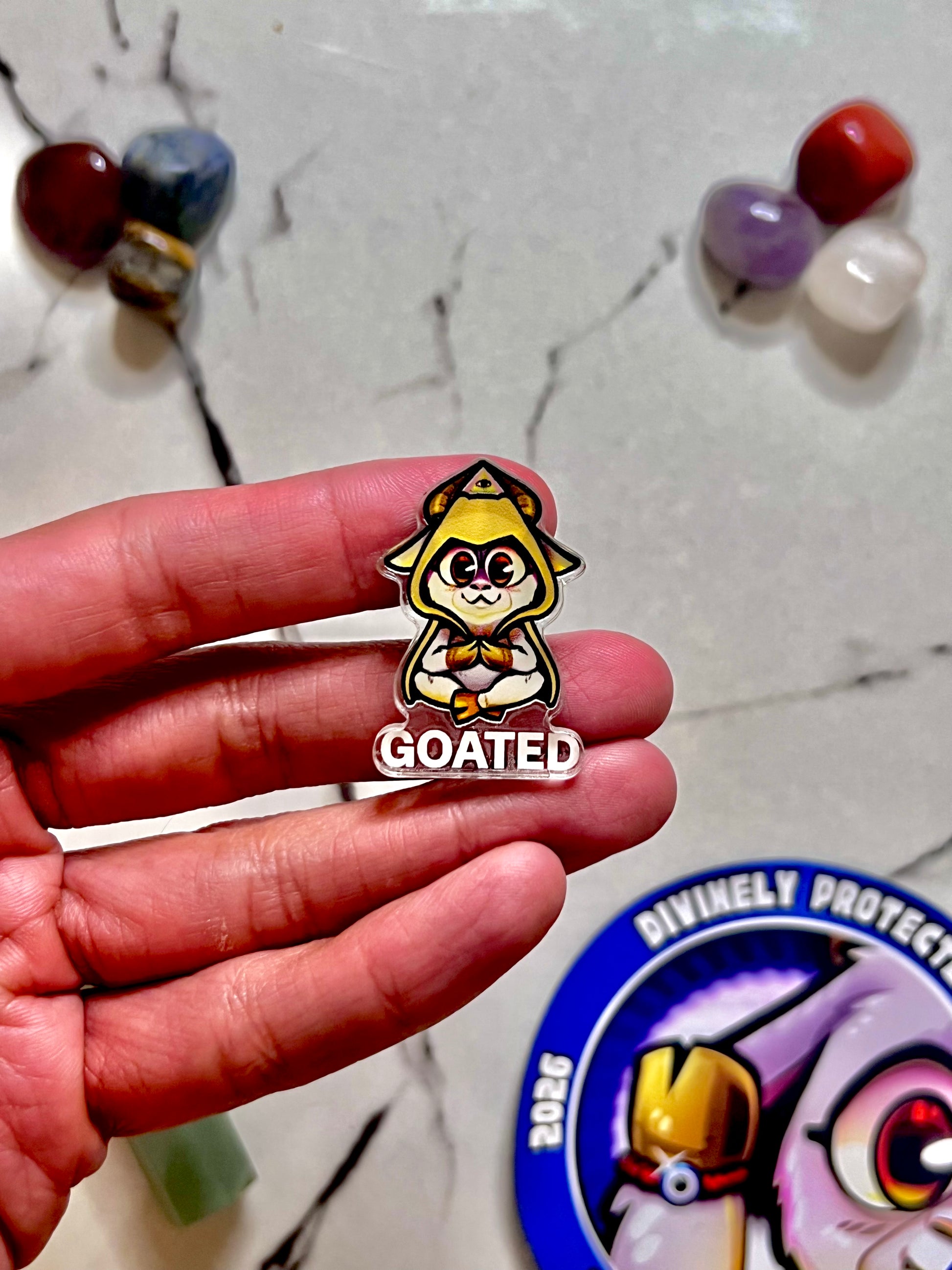 A hand holding a small enamel pin of a yellow cartoon goat character with the word 'GOATED' below it, set against a marble surface with blurred colorful stones and a circular pin backing partially visible.