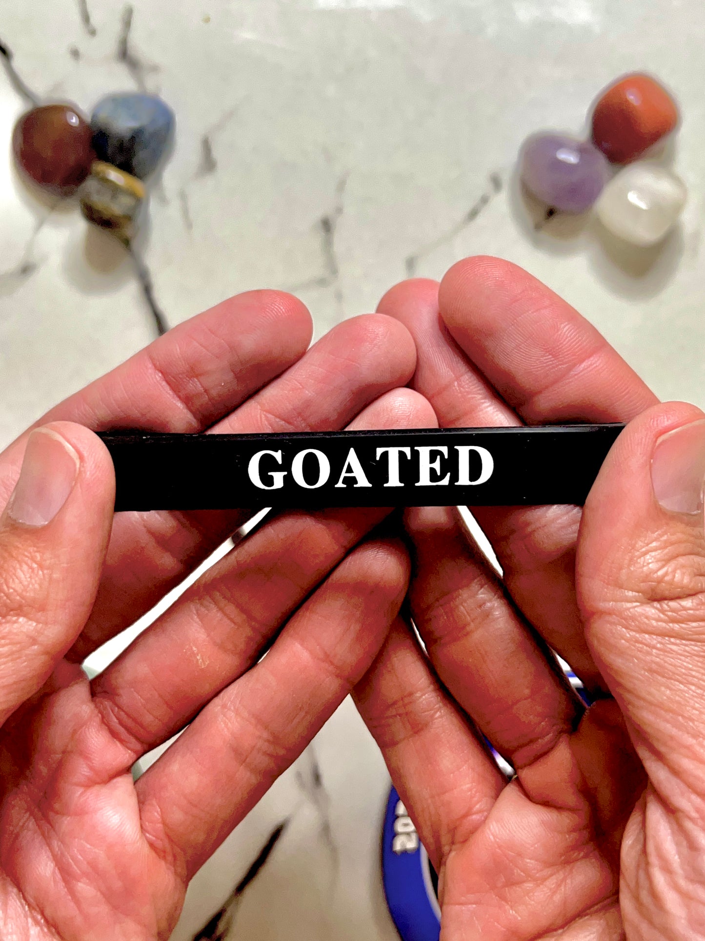 A person's hands holding a black silicone Bracelet wristband with the word 'GOATED' printed in white capital letters, set against a marble-like surface with blurred colorful stones in the background.