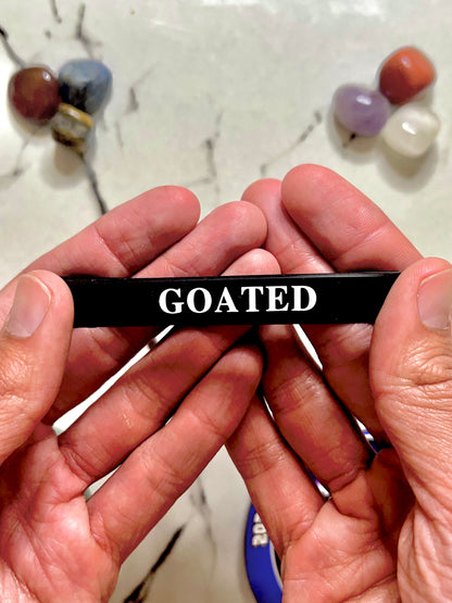 A person's hands holding a black silicone Bracelet wristband with the word 'GOATED' printed in white capital letters, set against a marble-like surface with blurred colorful stones in the background.