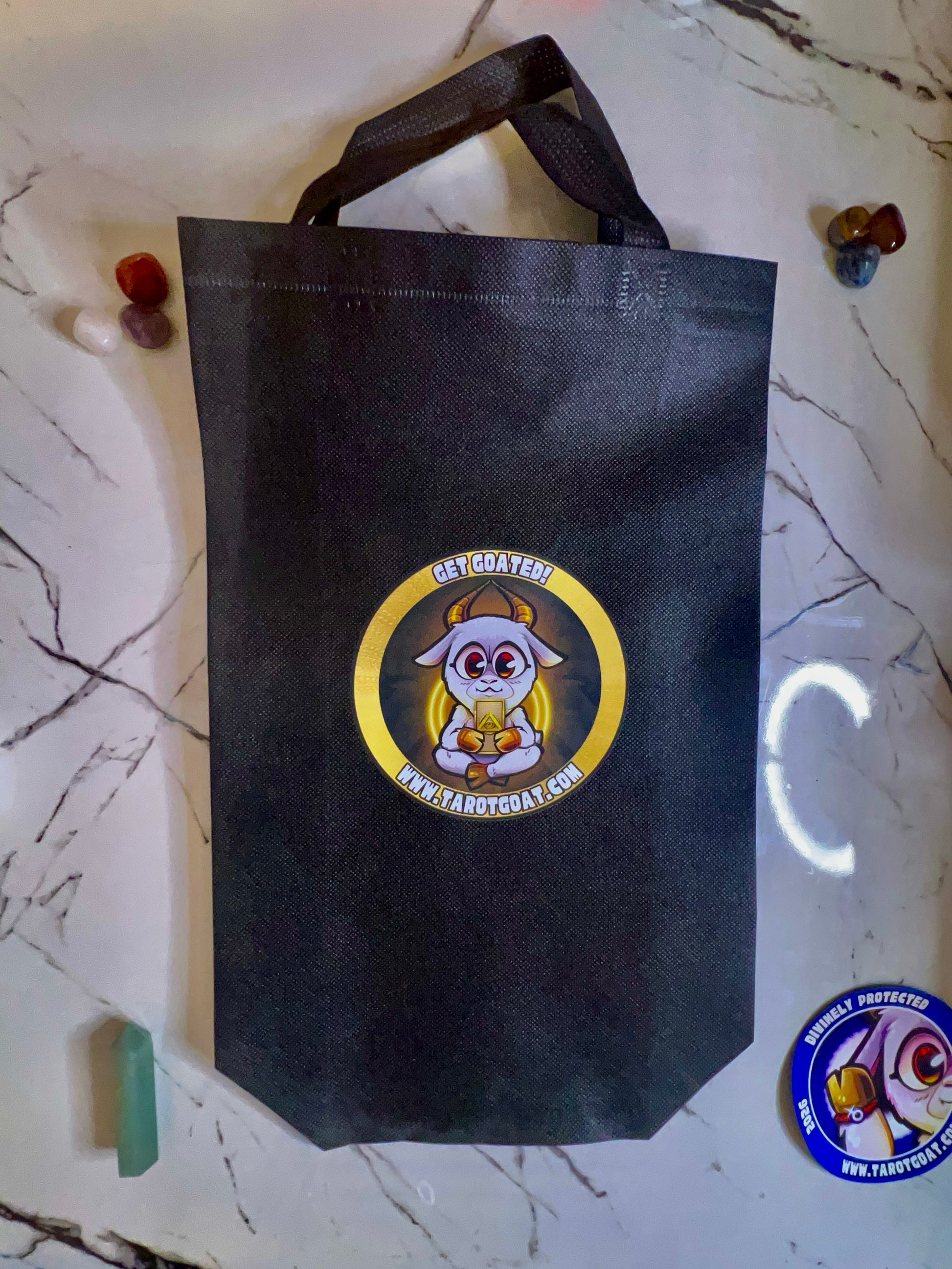 A black non-woven tote bag with a circular cartoon goat logo that reads 'GET GOATED' and 'WWW.TAROTGOAT.COM', placed on a white marble surface with small decorative stones around it.