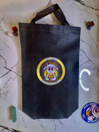 A black non-woven tote bag with a circular cartoon goat logo that reads 'GET GOATED' and 'WWW.TAROTGOAT.COM', placed on a white marble surface with small decorative stones around it.