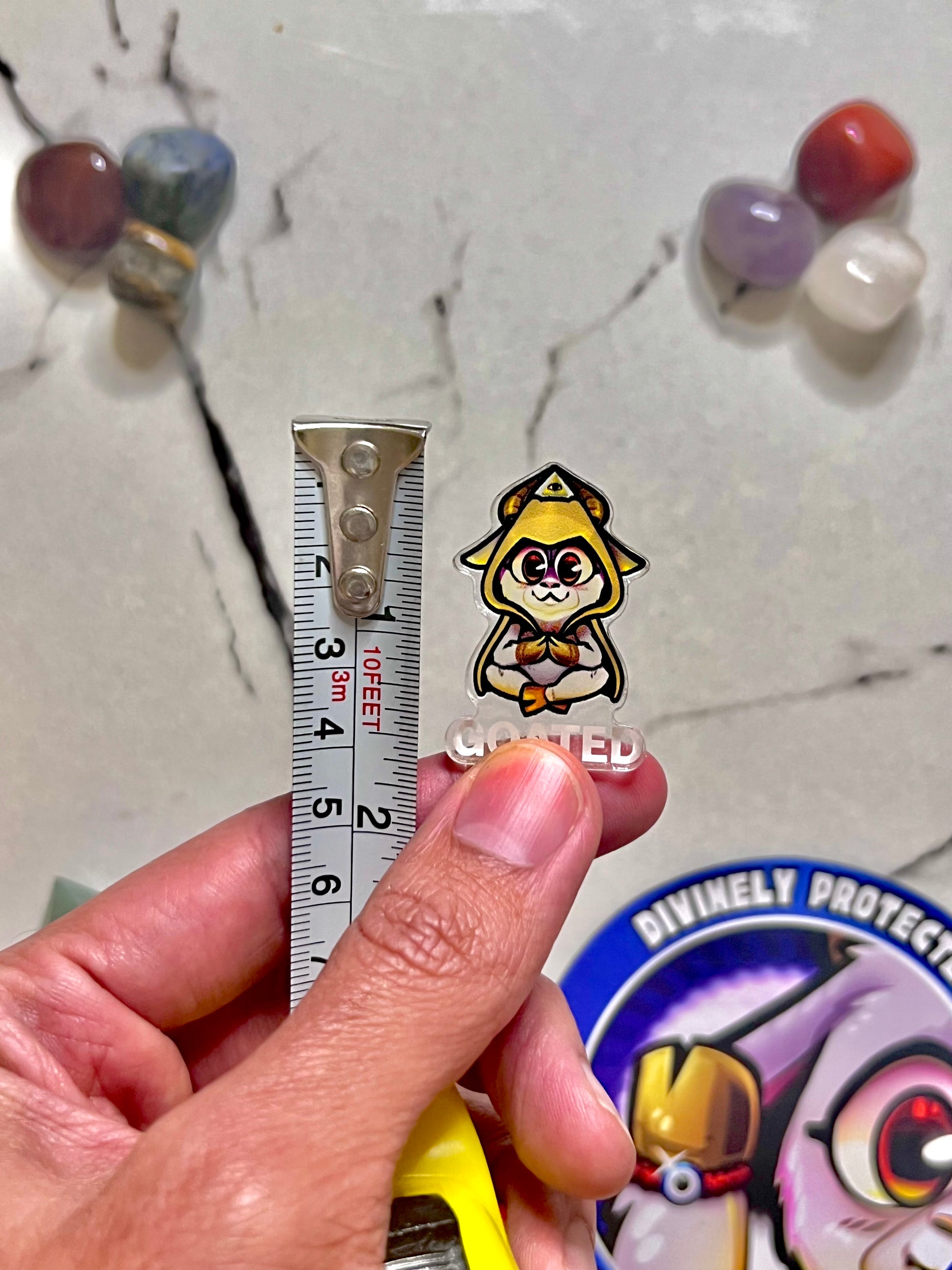 A hand holds a small anime-style enamel pin of a yellow-haired character with a red eye patch next to a metal ruler for scale, with a marble surface and colorful beads in the background.
