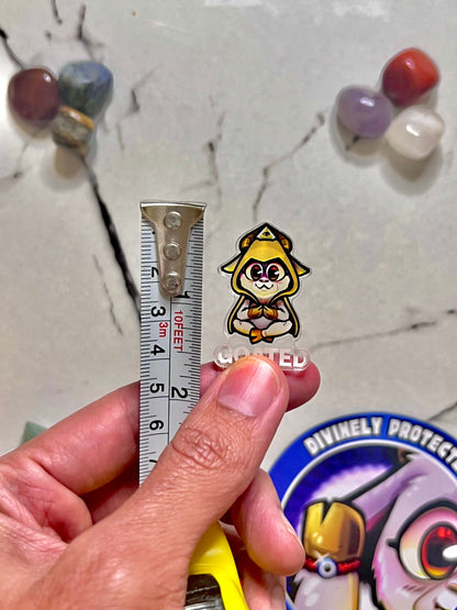 A hand holds a small anime-style enamel pin of a yellow-haired character with a red eye patch next to a metal ruler for scale, with a marble surface and colorful beads in the background.