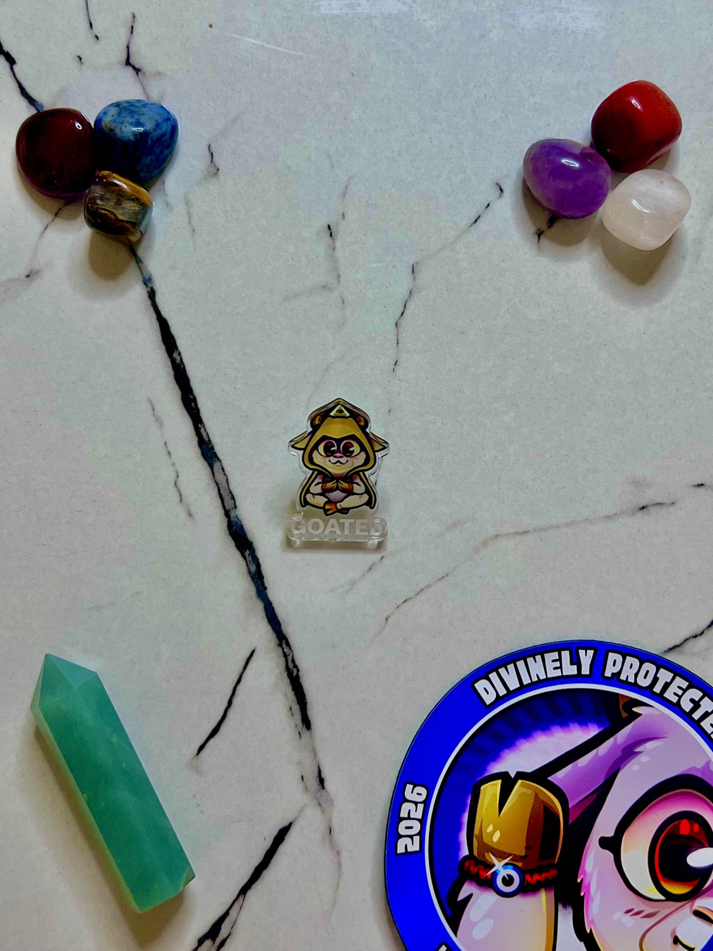 A small illustrated GOATED pin or charm featuring a Goat cartoon character in a yellow hat, placed on a white marble-like surface with crystals and a round sticker partially visible.
