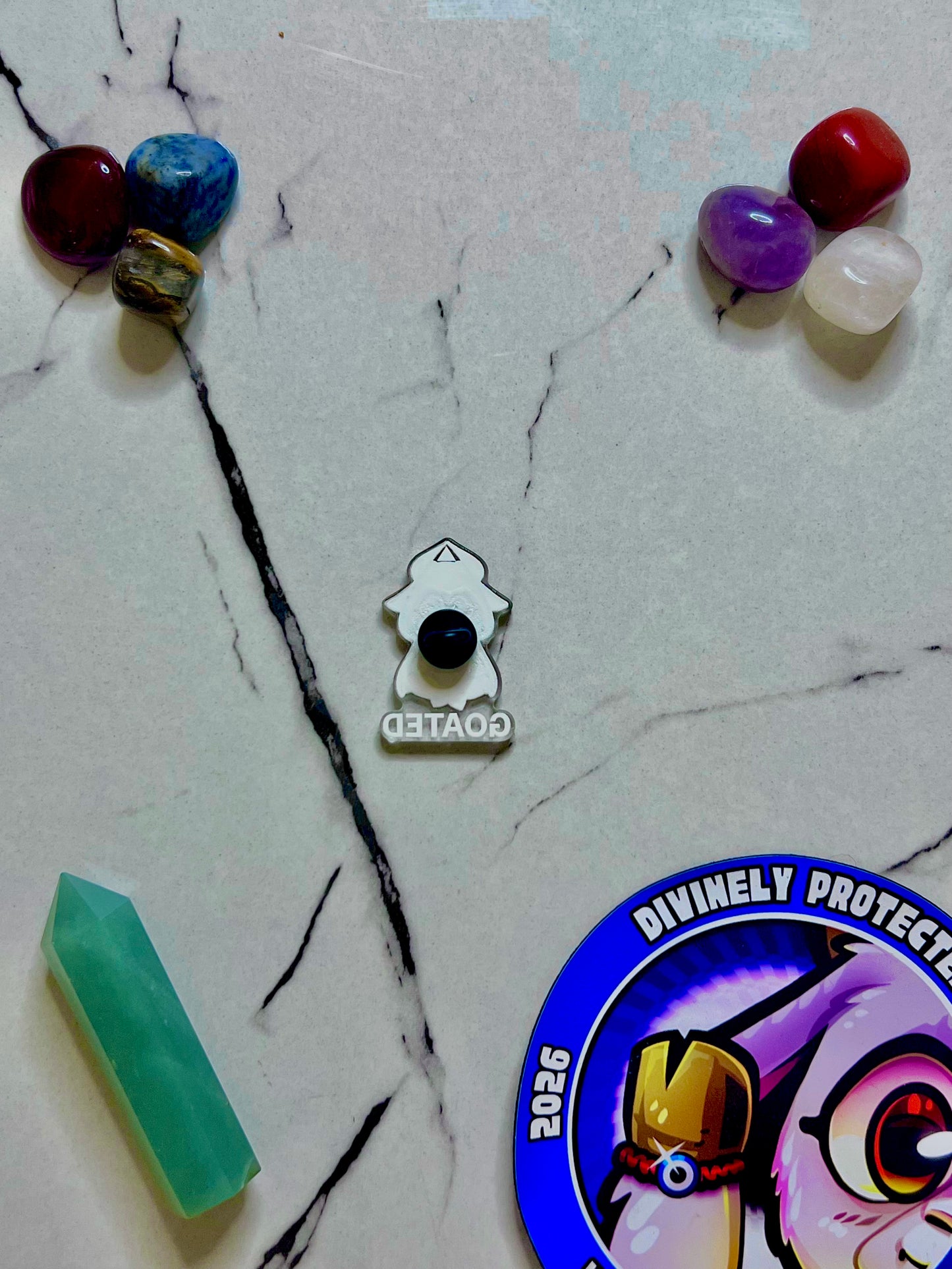 A small enamel pin shaped like a white owl with a black circular center and the word 'TETAOE' below, displayed on a light marble-like surface with crystals and a blue circular sticker around it.