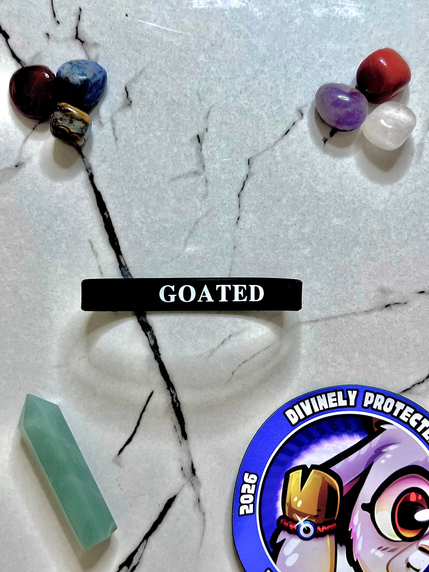 A black silicone wristband bracelet with the word 'GOATED' in white text, displayed on a white marble surface with various colorful crystals and a circular sticker.