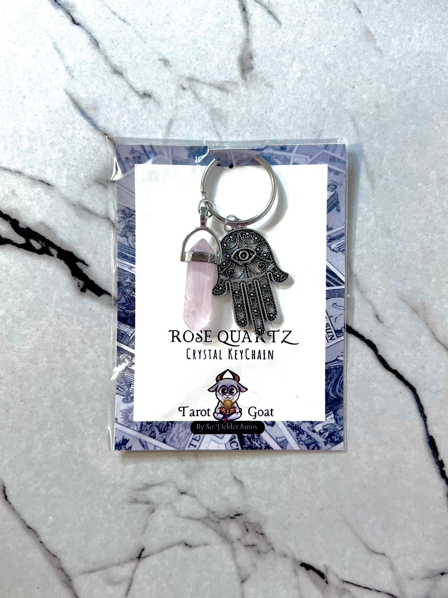 Rose Quartz crystal keychain with Hamsa hand charm on a branded card against a marble background