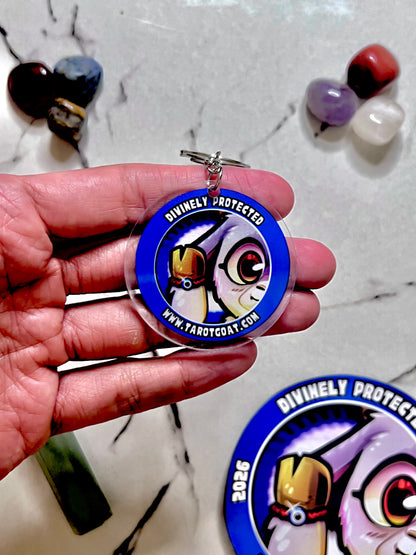 A hand holds a round acrylic keychain featuring a blue border and a goat cartoon owl illustration with the text 'Divinely Protected' and 'www.tarotcoat.