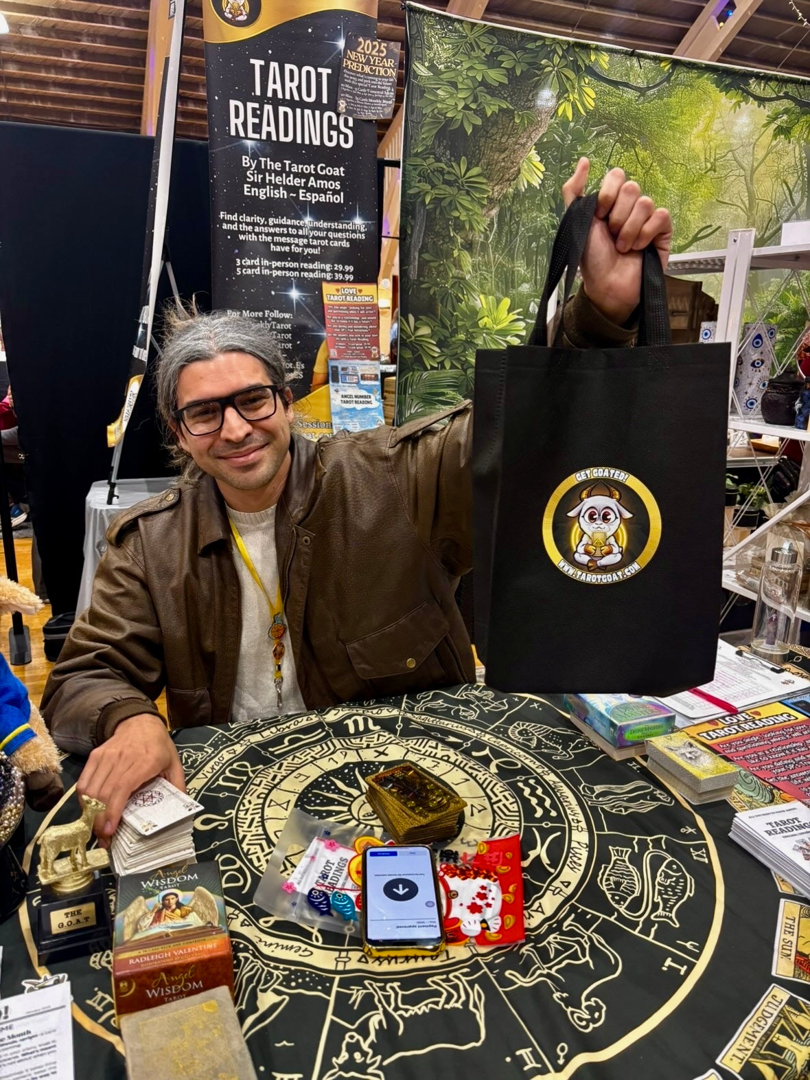 Sir Helder Amos at a booth, holds up a black tote bag with a circular Get Goated Tarot Goat logo. The booth displays tarot cards, a tarot reading sign, and other mystical items on a table.