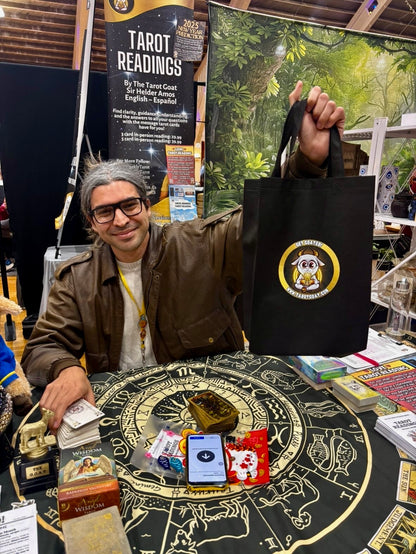 Sir Helder Amos at a booth, holds up a black tote bag with a circular Get Goated Tarot Goat logo. The booth displays tarot cards, a tarot reading sign, and other mystical items on a table.