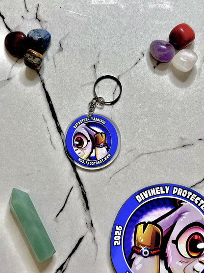 A circular acrylic keychain featuring a cartoon owl illustration with the text 'DIVINELY PROTEST' and '2026' on a blue background, displayed on a white marble surface with crystals and another partial matching keychain visible.