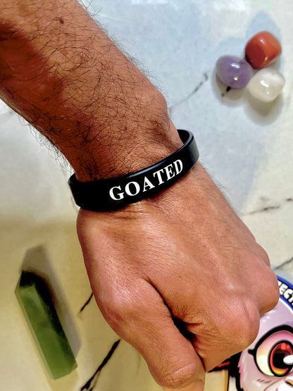 A close-up of a person's wrist wearing a black silicone bracelet with the word 'GOATED' printed in white capital letters. The background is a light marble-like surface with some decorative stones and a cartoon illustration partially visible at the bottom right.