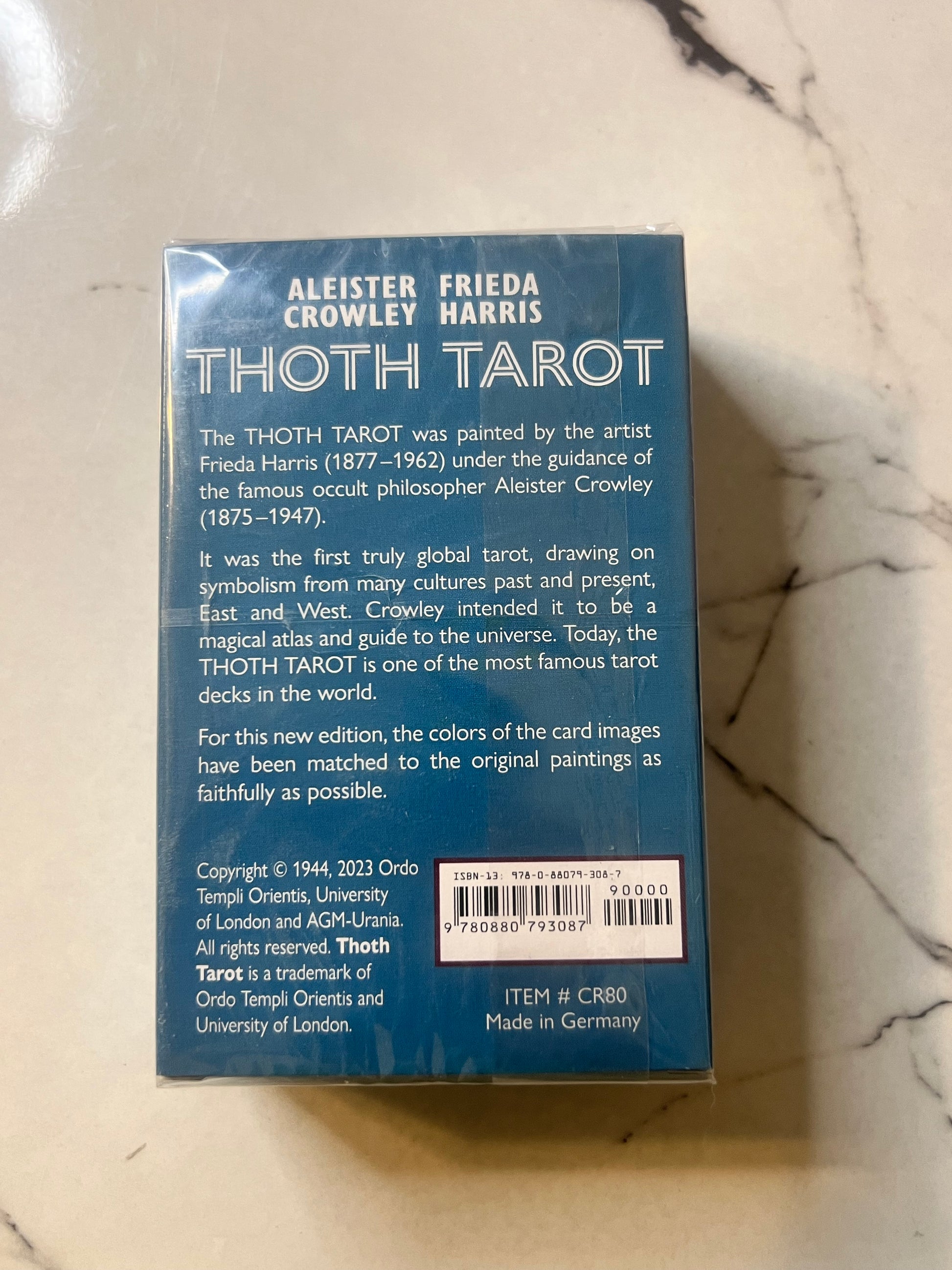 The back of the Thoth Tarot card deck box by Aleister Friedrich Crowley and Crowley Harris, placed on a white marble surface. The box is dark blue with white text detailing the book's history, card design, and publisher information by Ordo Templi Orientis.