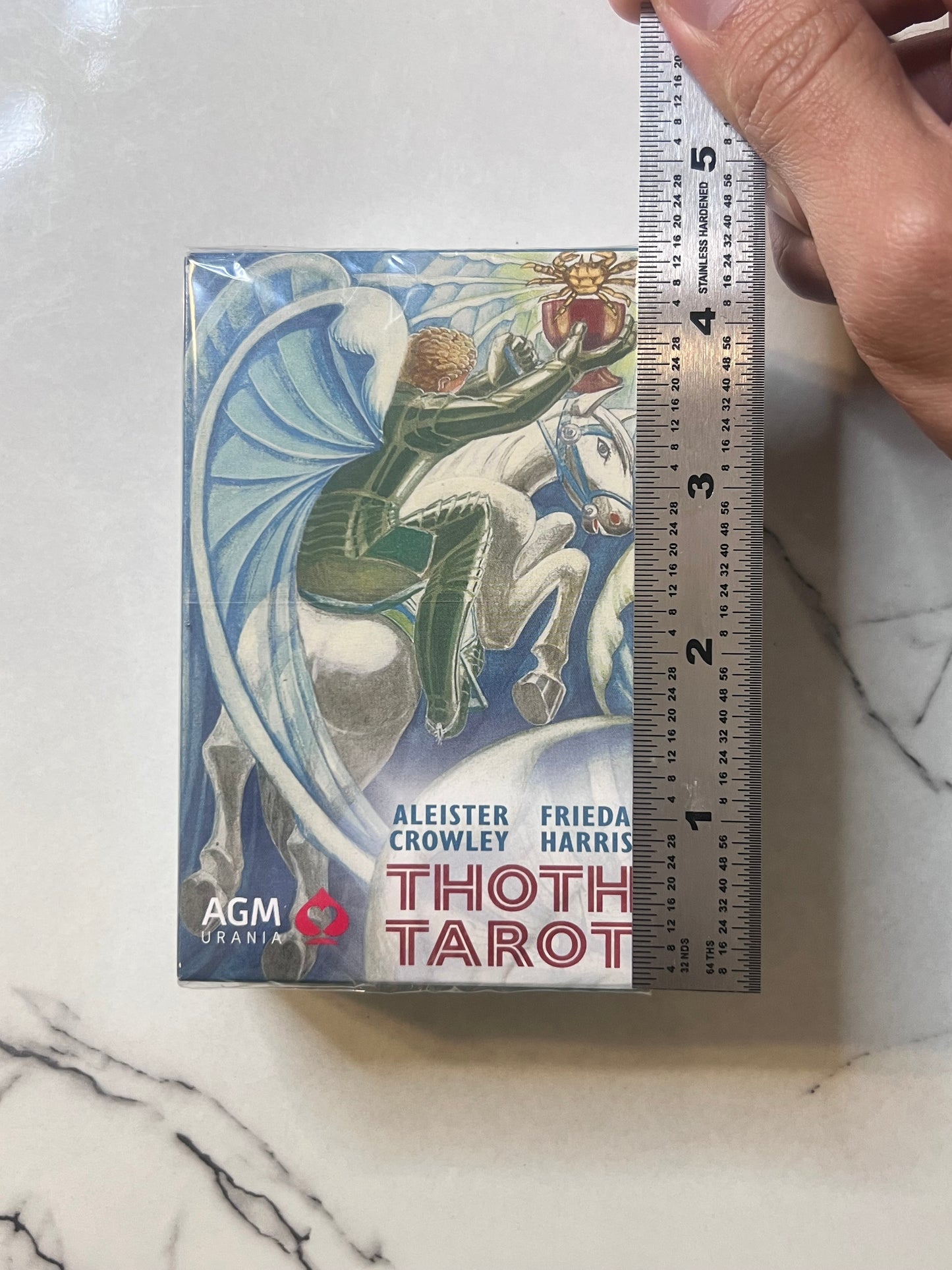 A boxed card deck labeled 'Thoth Tarot' with an illustrated winged rider on a white horse, placed on a marble-like surface next to a hand holding a ruler for scale. Visible text includes 'AGM Urania' and names Aleister Crowley and Frieda Harris.