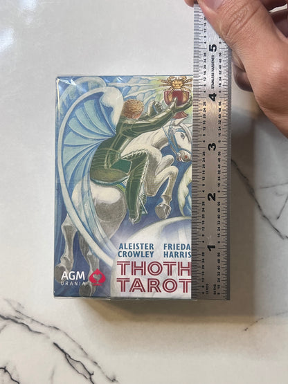 A boxed card deck labeled 'Thoth Tarot' with an illustrated winged rider on a white horse, placed on a marble-like surface next to a hand holding a ruler for scale. Visible text includes 'AGM Urania' and names Aleister Crowley and Frieda Harris.