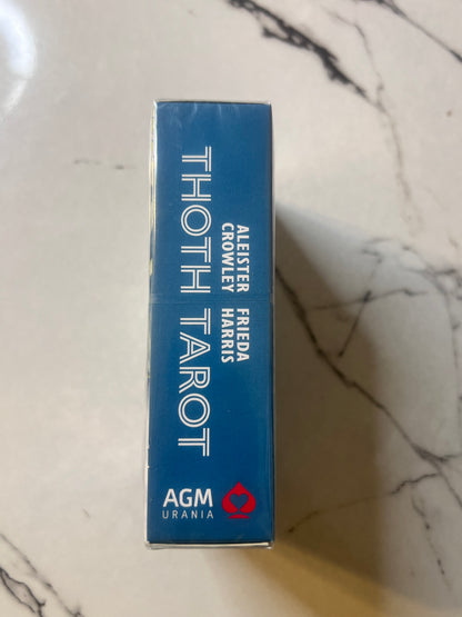 A blue rectangular box of AGM Urania Thoth Tarot standing upright on a white marble surface. The box features white text reading 'THOTH TAROT' and smaller text listing 'ALESTER FRIEDA CROWLEY HARKS'.